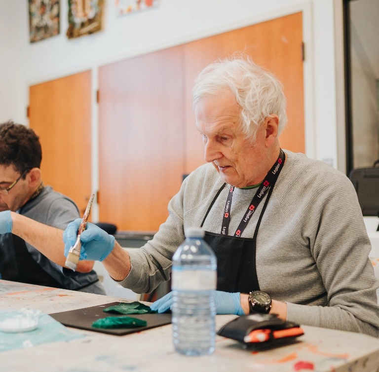 Grey haired man wearing a black apron and painting a picture at a table