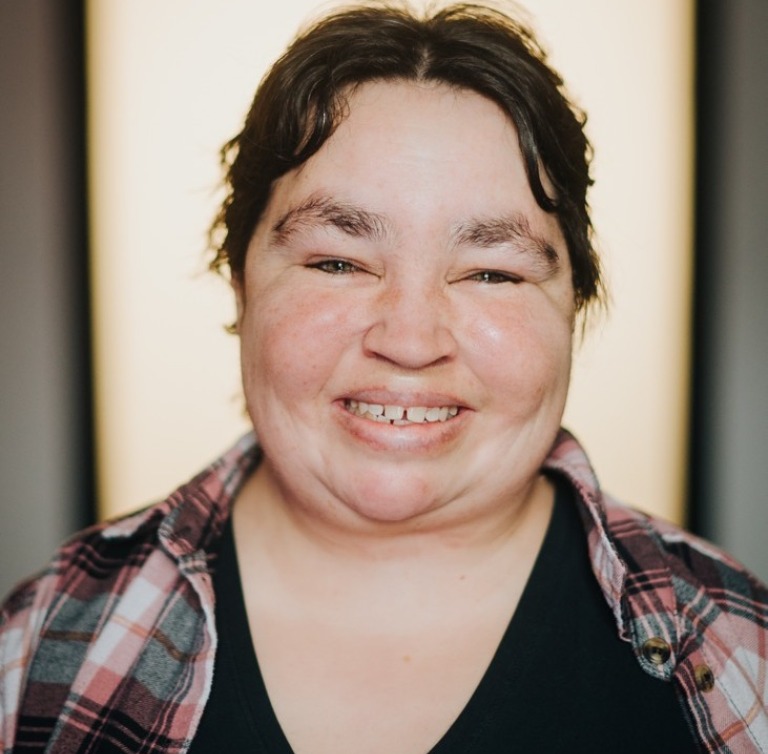 A woman with a round red face wearing a plaid shirt smiling happily