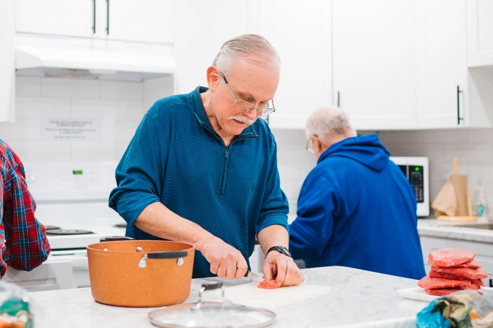 WHite haired man with a moustache in a blue top chops a carrot at a kitchen counter