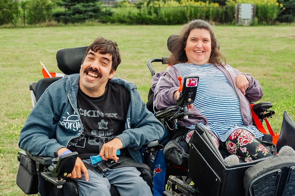 Dark haired man in a black shirt and a long brown haired woman in a striped top smile from their wheels chairs outside
